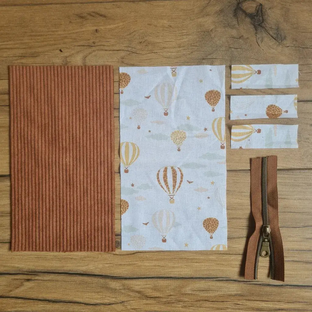 Kit "prêt à coudre" pour une trousse berlingot en velours côtelé au coloris marron avec une doublure en tissu à motif montgolfières. L'ensemble des pièces de tissu, déjà prédécoupées aux bonnes dimensions, sont disposées au sol. A gauche, une pièce en velours côtelé marron. A droite, quatre pièces en tissu imprimé à motif montgolfières ainsi qu'une fermeture éclair marron.
