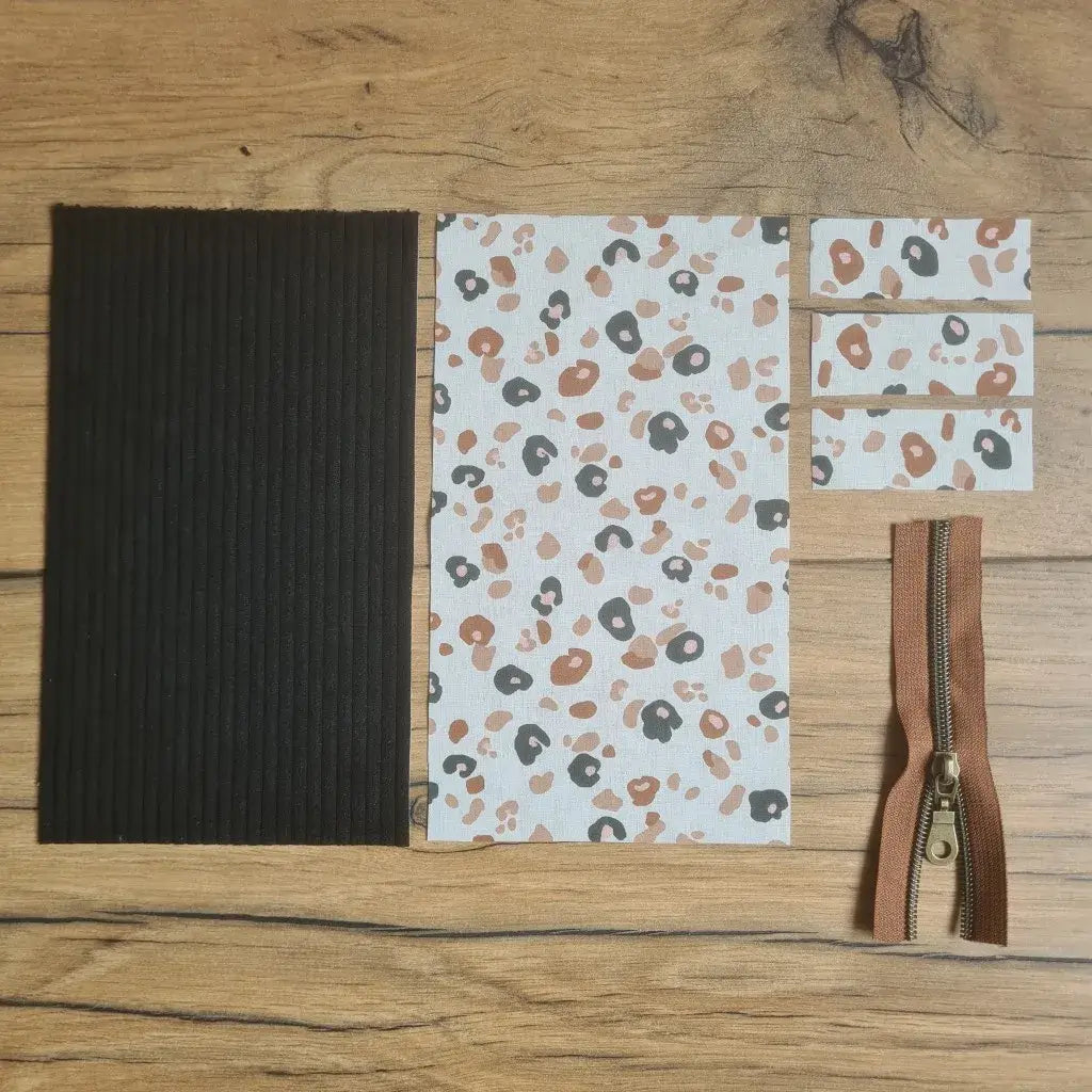 Kit "prêt à coudre" pour une trousse berlingot en velours côtelé au coloris noir avec une doublure en tissu à motif léopard. L'ensemble des pièces de tissu, déjà prédécoupées aux bonnes dimensions, sont disposées au sol. A gauche, une pièce en velours côtelé noir. A droite, quatre pièces en tissu imprimé à motif léopard ainsi qu'une fermeture éclair marron.