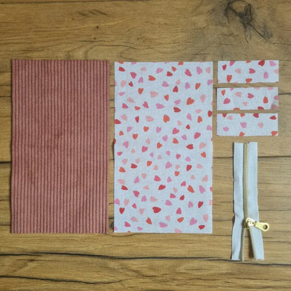 Kit "prêt à coudre" pour une trousse berlingot en velours côtelé au coloris rose avec une doublure en tissu à motif cœurs. L'ensemble des pièces de tissu, déjà prédécoupées aux bonnes dimensions, sont disposées au sol. A gauche, une pièce en velours côtelé rose. A droite, quatre pièces en tissu imprimé à motif cœurs ainsi qu'une fermeture éclair blanche.