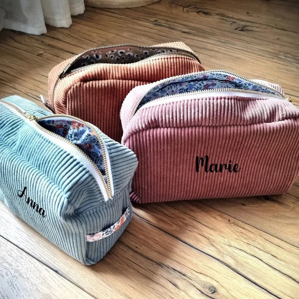 Trois trousses de toilette en velours côtelé disposées au sol sur un parquet en chêne. La trousse de droite est de coloris bleu ciel avec un tissu intérieur à motif fleuri est personnalisée au prénom "Anna". Celle de droite est en velours côtelé rose avec un tissu intérieur à motif fleuri et est personnalisée au prénom "Marie". Celle en arrière plan est en velours côtelé marron avec un tissu intérieur au motif léopard.