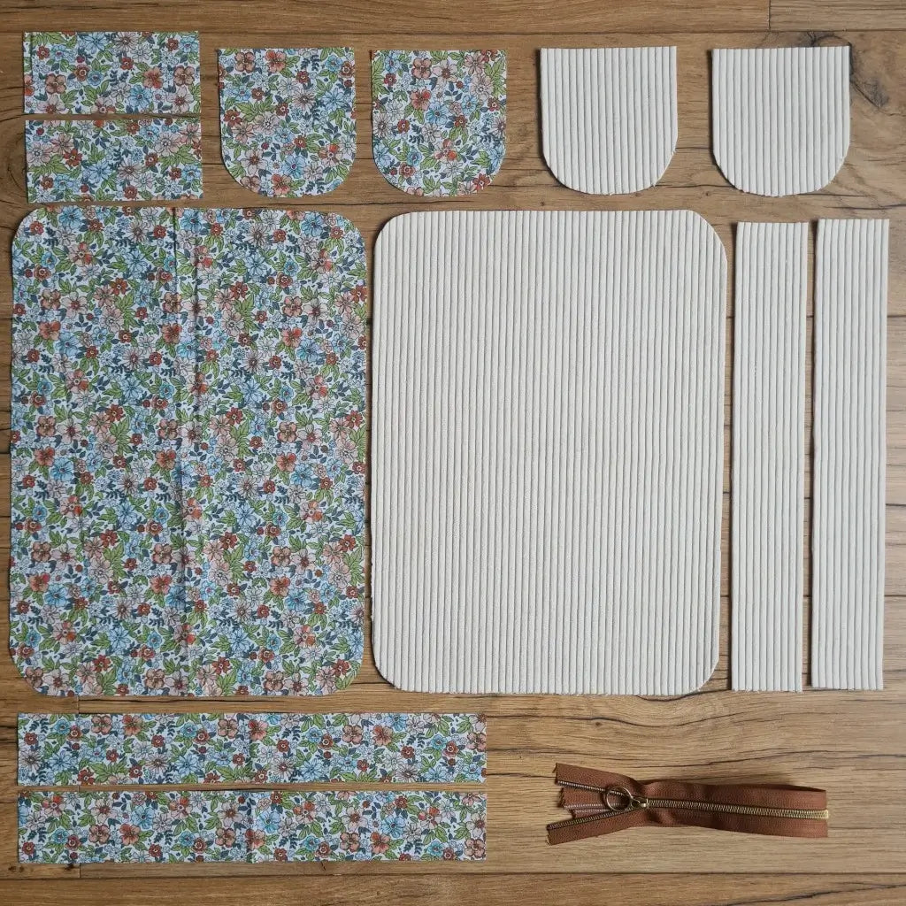 Kit "prêt à coudre" pour une trousse de toilette en velours côtelé au coloris beige avec une doublure en tissu à motif fleuri. L'ensemble des pièces de tissu, déjà prédécoupées aux bonnes dimensions, sont disposées au sol. A gauche, sept pièces en tissu à motif fleuri. A droite, cinq pièces en velours côtelé de couleur beige ainsi qu'une fermeture éclair marron.