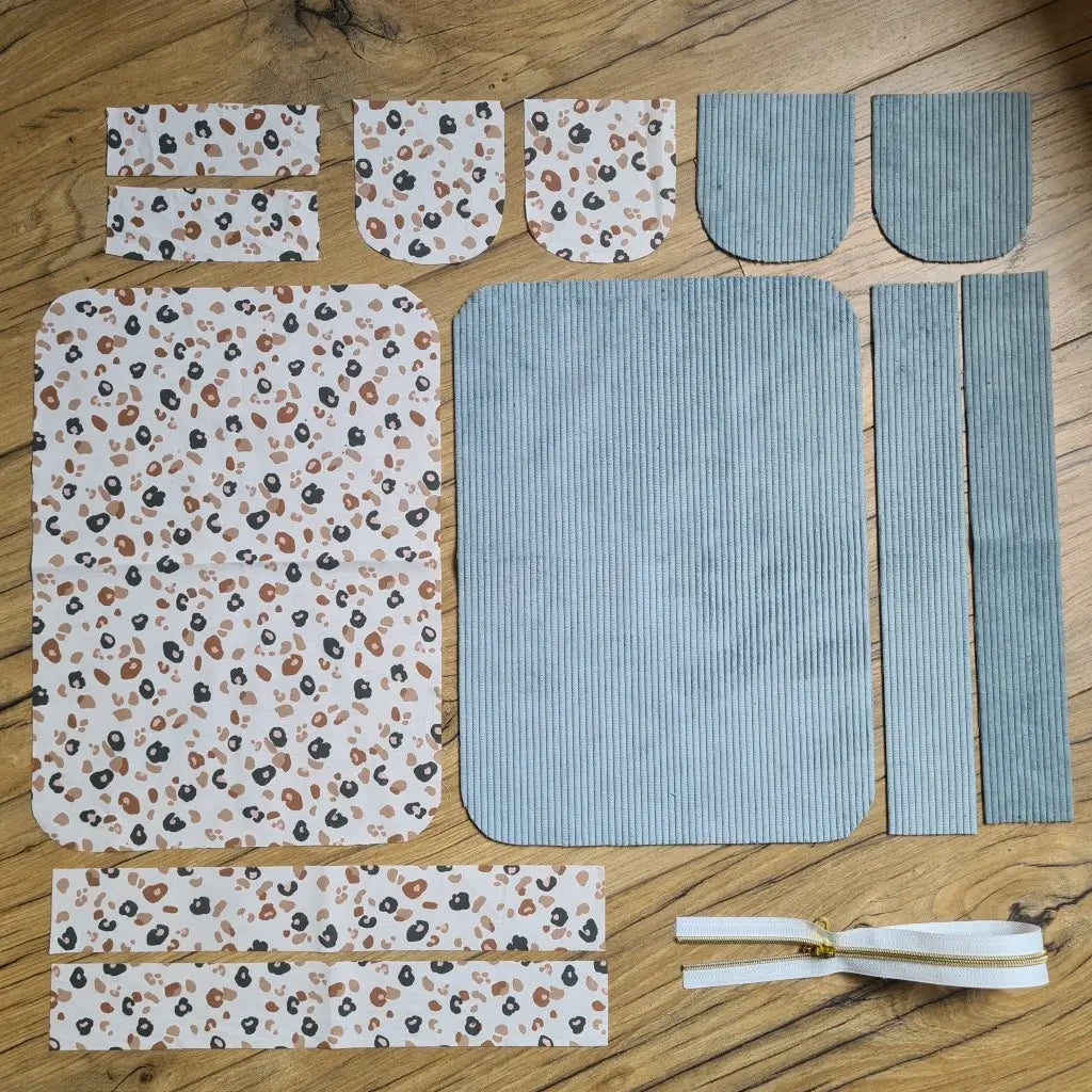 Kit "prêt à coudre" pour une trousse de toilette en velours côtelé au coloris bleu ciel avec une doublure en tissu à motif léopard. L'ensemble des pièces de tissu, déjà prédécoupées aux bonnes dimensions, sont disposées au sol. A gauche, sept pièces en tissu à motif léopard. A droite, cinq pièces en velours côtelé de couleur bleu ciel ainsi qu'une fermeture éclair blanche.