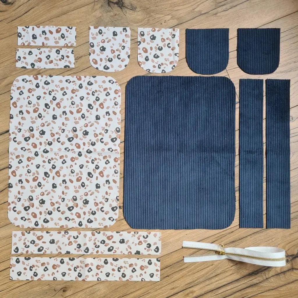 Kit "prêt à coudre" pour une trousse de toilette en velours côtelé au coloris bleu foncé avec une doublure en tissu à motif léopard. L'ensemble des pièces de tissu, déjà prédécoupées aux bonnes dimensions, sont disposées au sol. A gauche, sept pièces en tissu à motif léopard. A droite, cinq pièces en velours côtelé de couleur bleu foncé ainsi qu'une fermeture éclair blanche.