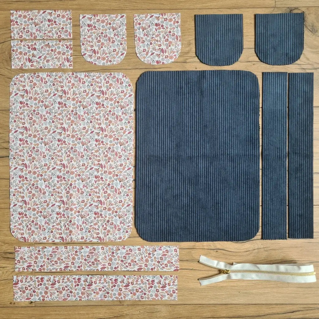 Kit "prêt à coudre" pour une trousse de toilette en velours côtelé au coloris bleu foncé avec une doublure en tissu à motif liberty. L'ensemble des pièces de tissu, déjà prédécoupées aux bonnes dimensions, sont disposées au sol. A gauche, sept pièces en tissu à motif liberty. A droite, cinq pièces en velours côtelé de couleur bleu foncé ainsi qu'une fermeture éclair blanche.