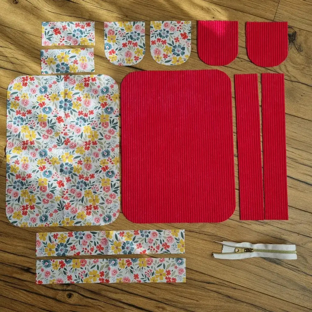 Ensemble de pièces de tissu prêtes à assembler pour confectionner une trousse de toilette. Les éléments comprennent du velours côtelé framboise uni et un tissu imprimé floral multicolore aux tons jaune, bleu, rouge et rose, disposés à plat sur une table en bois. On distingue également une fermeture éclair blanche et dorée. Ce kit de couture permet de réaliser une création artisanale unique et personnalisée.