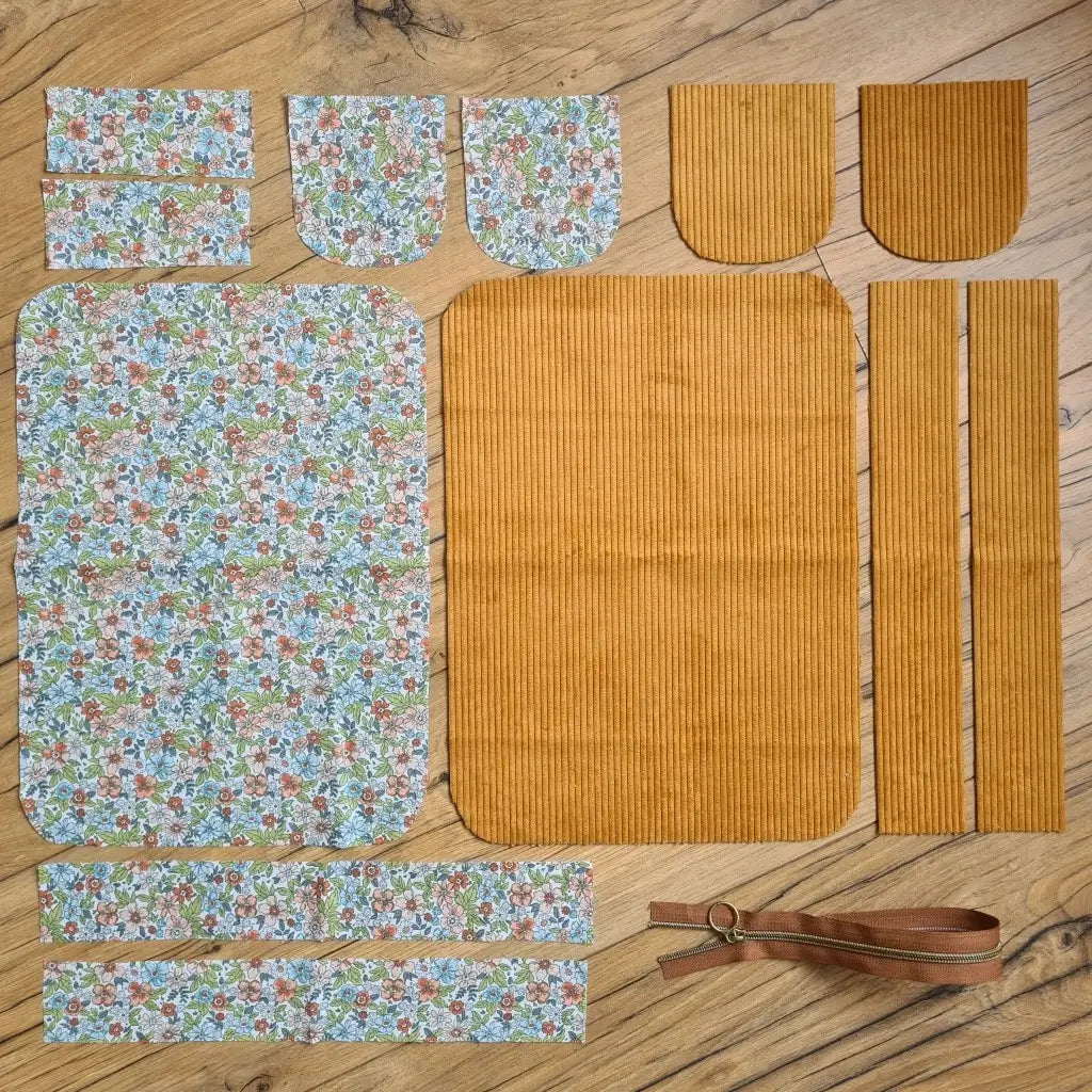 Kit "prêt à coudre" pour une trousse de toilette en velours côtelé au coloris jaune avec une doublure en tissu à motif fleuri. L'ensemble des pièces de tissu, déjà prédécoupées aux bonnes dimensions, sont disposées au sol. A gauche, sept pièces en tissu à motif fleuri. A droite, cinq pièces en velours côtelé de couleur jaune ainsi qu'une fermeture éclair marron.