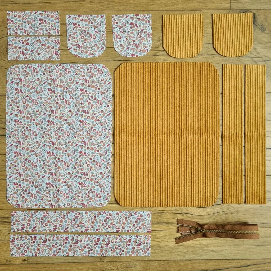 Kit "prêt à coudre" pour une trousse de toilette en velours côtelé au coloris jaune avec une doublure en tissu à motif liberty. L'ensemble des pièces de tissu, déjà prédécoupées aux bonnes dimensions, sont disposées au sol. A gauche, sept pièces en tissu à motif liberty. A droite, cinq pièces en velours côtelé de couleur jaune ainsi qu'une fermeture éclair marron.