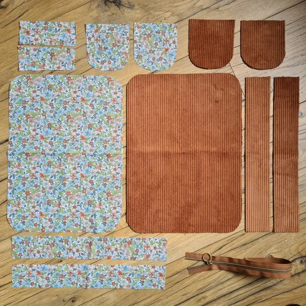 Kit "prêt à coudre" pour une trousse de toilette en velours côtelé au coloris marron avec une doublure en tissu à motif fleuri. L'ensemble des pièces de tissu, déjà prédécoupées aux bonnes dimensions, sont disposées au sol. A gauche, sept pièces en tissu à motif fleuri. A droite, cinq pièces en velours côtelé de couleur marron ainsi qu'une fermeture éclair marron.