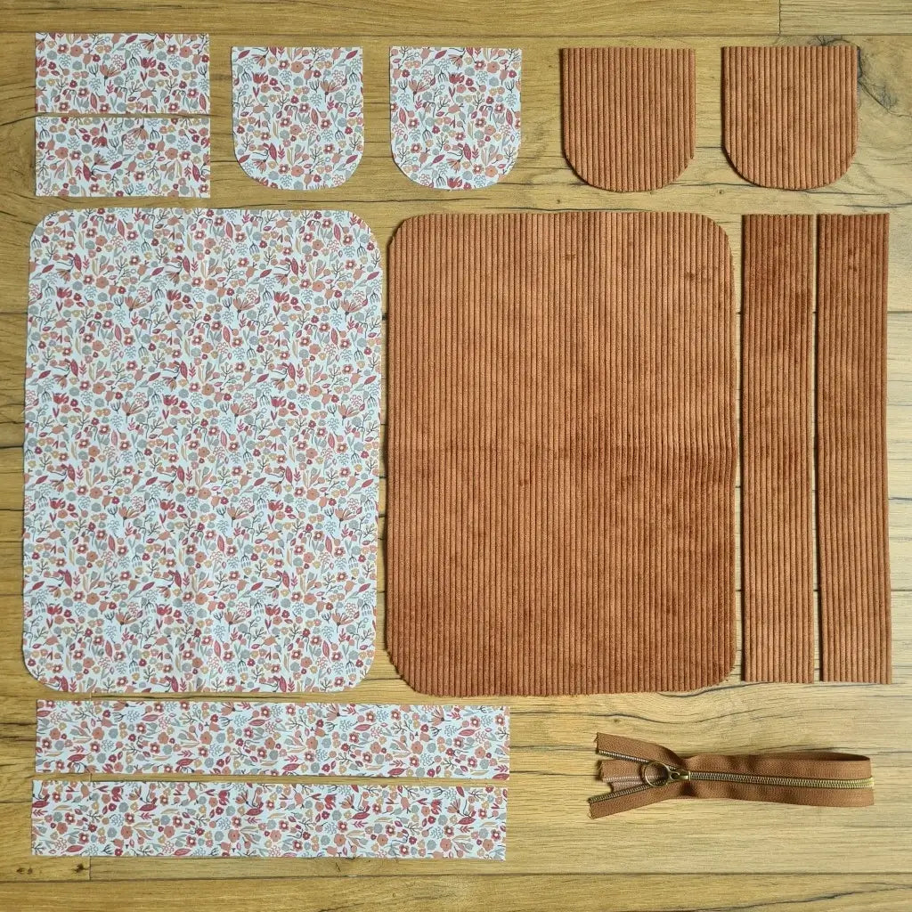 Kit "prêt à coudre" pour une trousse de toilette en velours côtelé au coloris marron avec une doublure en tissu à motif liberty. L'ensemble des pièces de tissu, déjà prédécoupées aux bonnes dimensions, sont disposées au sol. A gauche, sept pièces en tissu à motif liberty. A droite, cinq pièces en velours côtelé de couleur marron ainsi qu'une fermeture éclair marron.