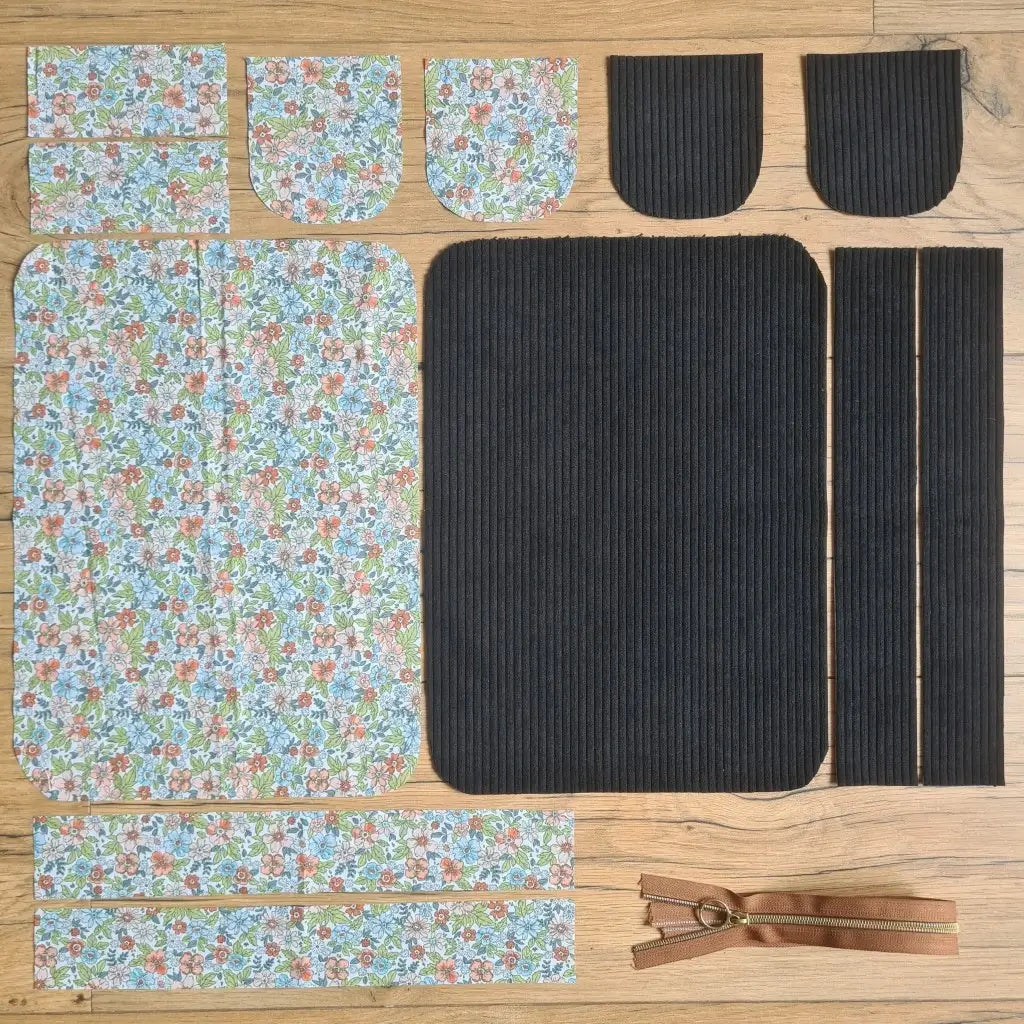 Kit "prêt à coudre" pour une trousse de toilette en velours côtelé au coloris noir avec une doublure en tissu à motif fleuri. L'ensemble des pièces de tissu, déjà prédécoupées aux bonnes dimensions, sont disposées au sol. A gauche, sept pièces en tissu à motif fleuri. A droite, cinq pièces en velours côtelé de couleur noire ainsi qu'une fermeture éclair marron.