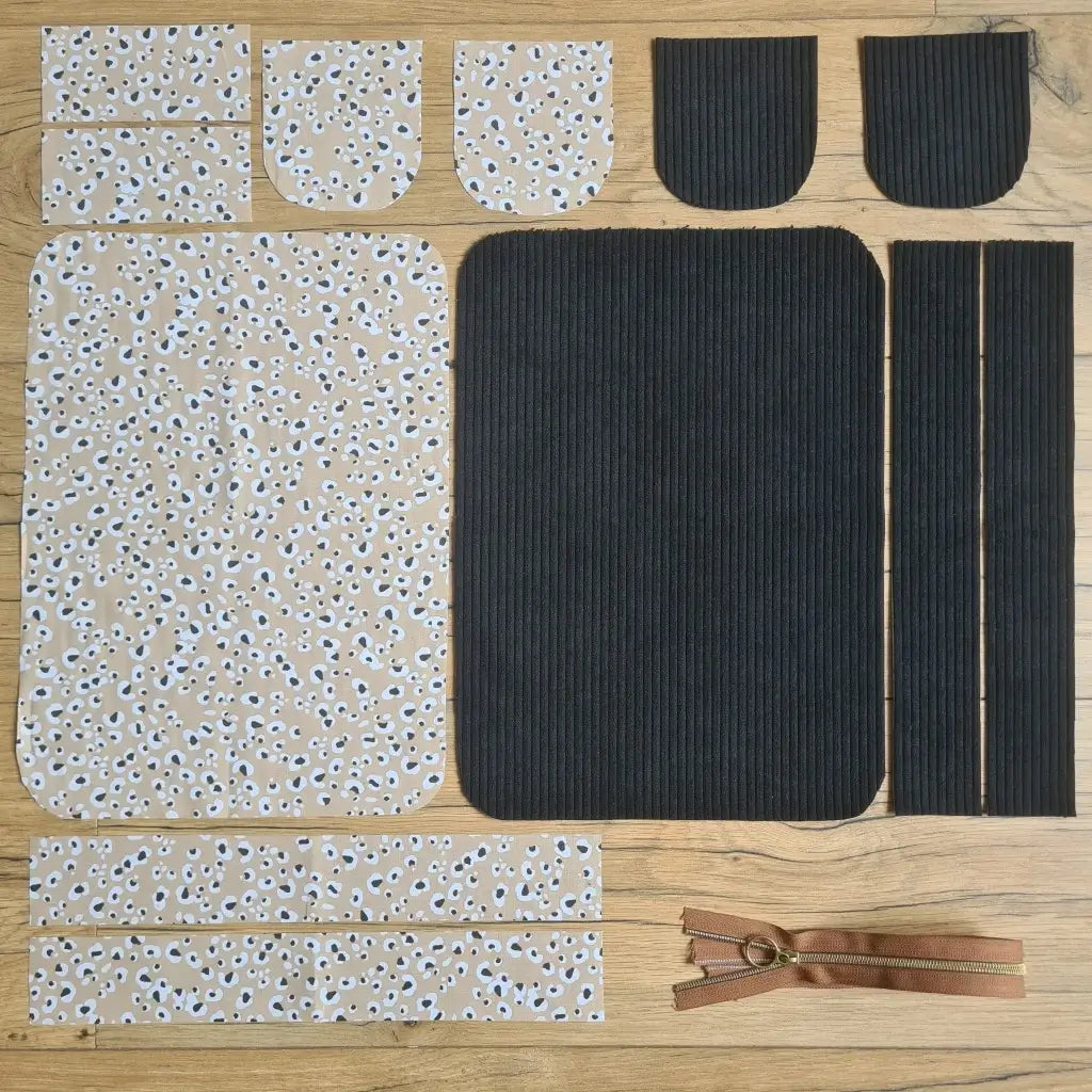 Kit "prêt à coudre" pour une trousse de toilette en velours côtelé au coloris noir avec une doublure en tissu à motif léopard. L'ensemble des pièces de tissu, déjà prédécoupées aux bonnes dimensions, sont disposées au sol. A gauche, sept pièces en tissu à motif léopard. A droite, cinq pièces en velours côtelé de couleur noire ainsi qu'une fermeture éclair marron.