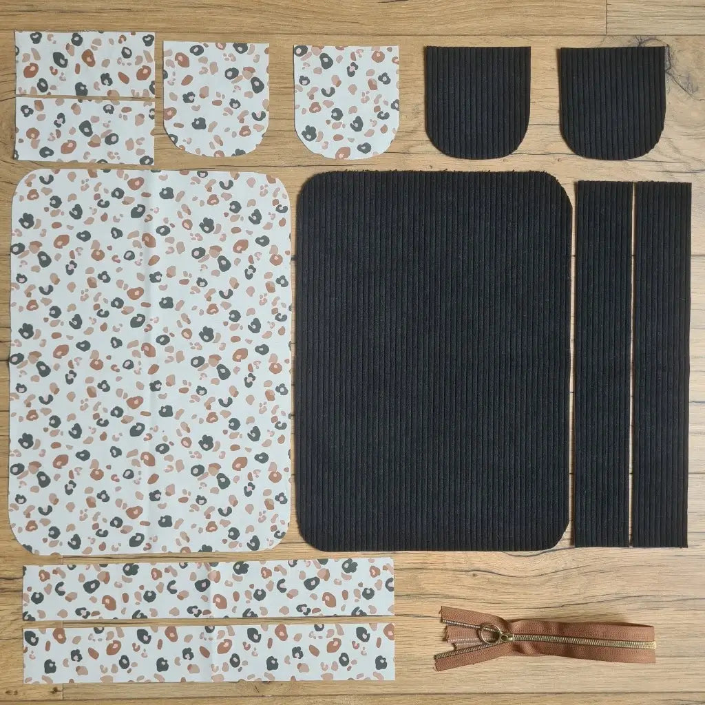 Kit "prêt à coudre" pour une trousse de toilette en velours côtelé au coloris noir avec une doublure en tissu à motif léopard. L'ensemble des pièces de tissu, déjà prédécoupées aux bonnes dimensions, sont disposées au sol. A gauche, sept pièces en tissu à motif léopard. A droite, cinq pièces en velours côtelé de couleur noire ainsi qu'une fermeture éclair marron.