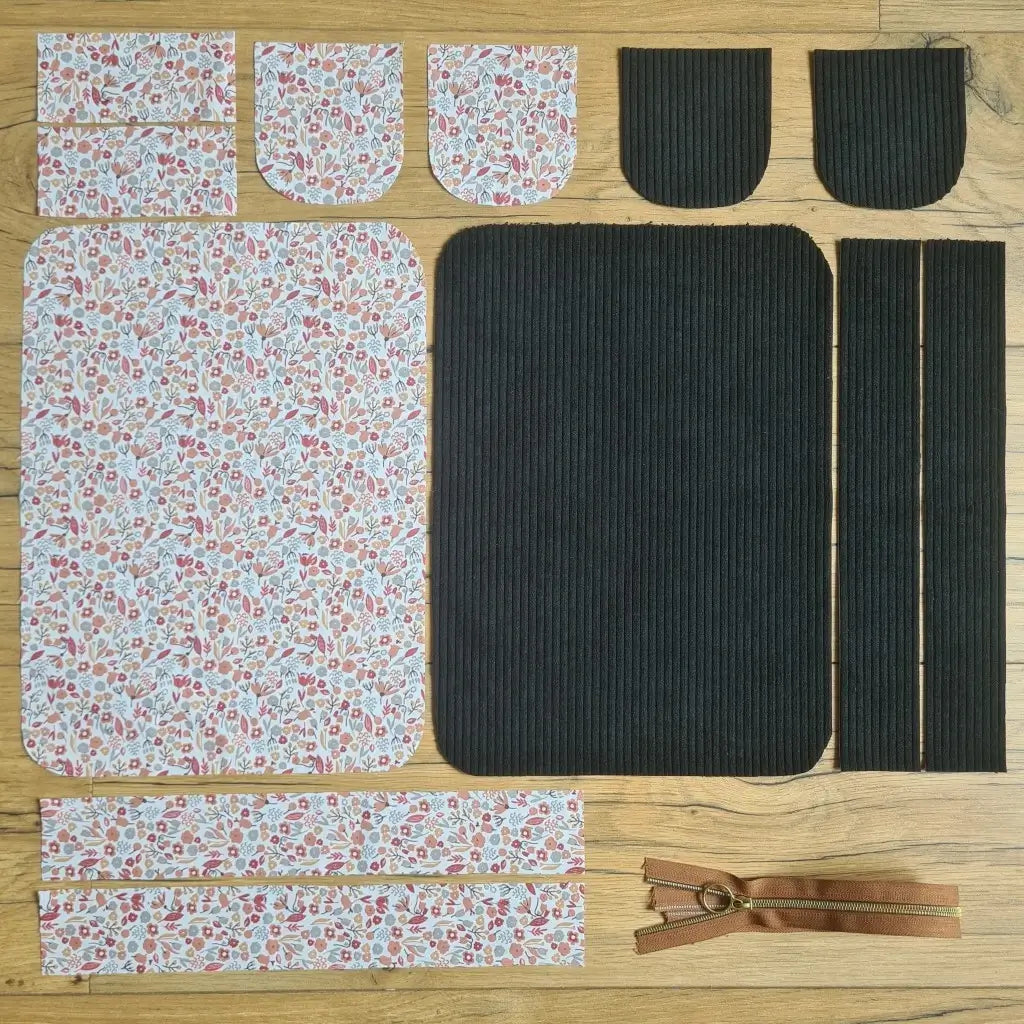 Kit "prêt à coudre" pour une trousse de toilette en velours côtelé au coloris noir avec une doublure en tissu à motif liberty. L'ensemble des pièces de tissu, déjà prédécoupées aux bonnes dimensions, sont disposées au sol. A gauche, sept pièces en tissu à motif liberty. A droite, cinq pièces en velours côtelé de couleur noire ainsi qu'une fermeture éclair marron.