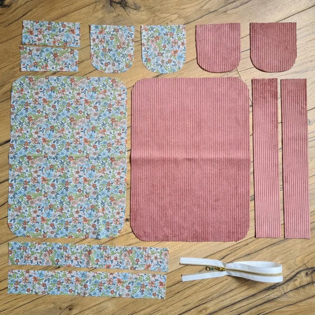 Kit "prêt à coudre" pour une trousse de toilette en velours côtelé au coloris rose avec une doublure en tissu à motif fleuri. L'ensemble des pièces de tissu, déjà prédécoupées aux bonnes dimensions, sont disposées au sol. A gauche, sept pièces en tissu à motif fleuri. A droite, cinq pièces en velours côtelé de couleur rose ainsi qu'une fermeture éclair blanche.