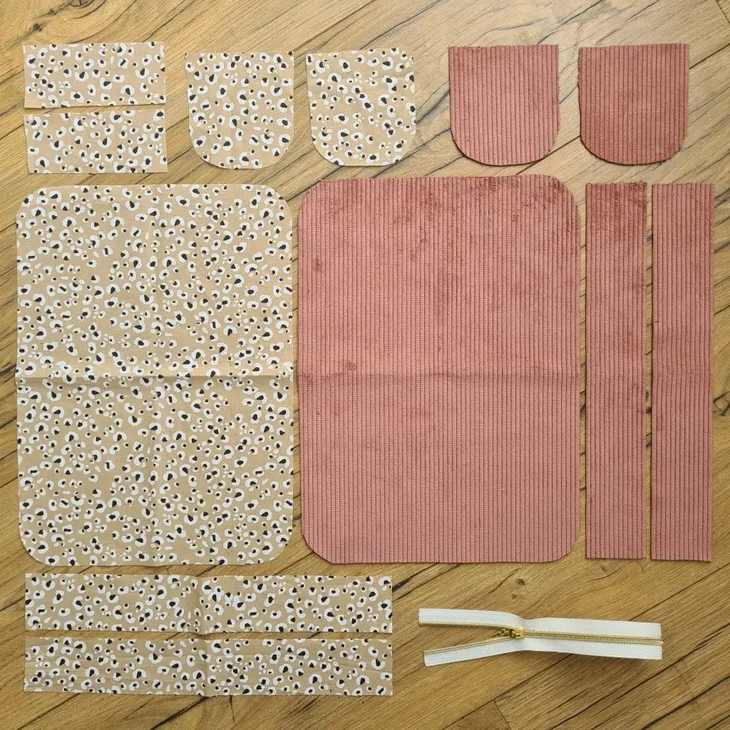 Kit "prêt à coudre" pour une trousse de toilette en velours côtelé au coloris rose avec une doublure en tissu à motif léopard. L'ensemble des pièces de tissu, déjà prédécoupées aux bonnes dimensions, sont disposées au sol. A gauche, sept pièces en tissu à motif léopard. A droite, cinq pièces en velours côtelé de couleur rose ainsi qu'une fermeture éclair blanche.