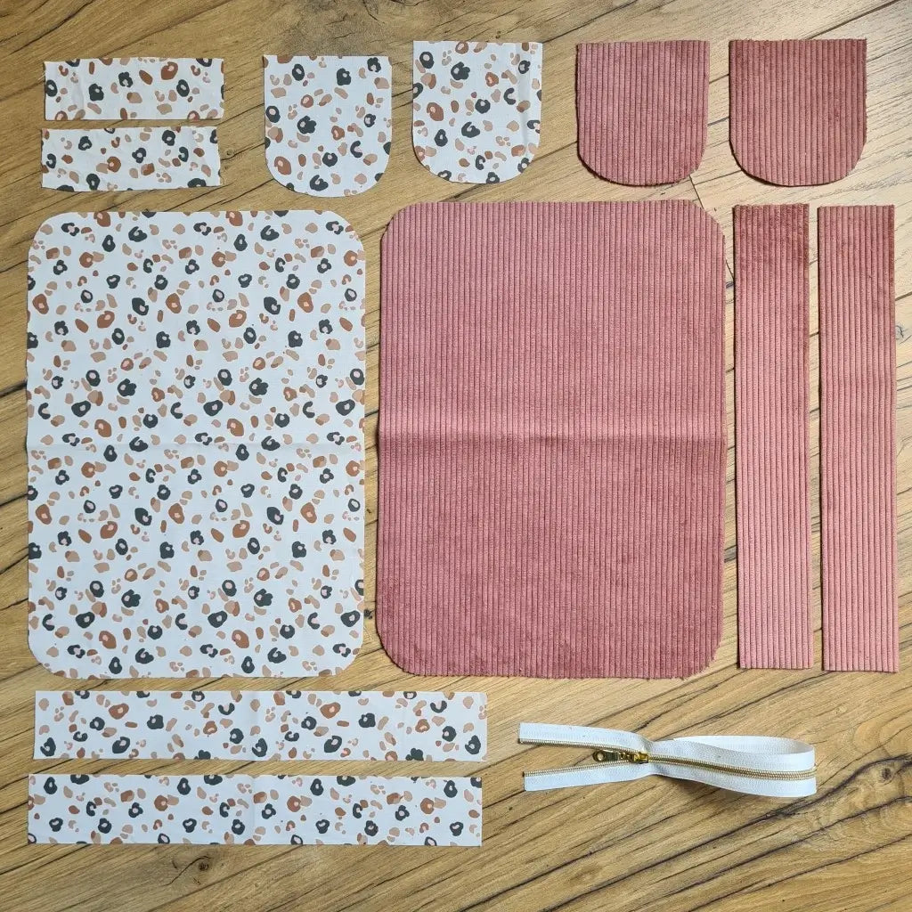 Kit "prêt à coudre" pour une trousse de toilette en velours côtelé au coloris rose avec une doublure en tissu à motif léopard. L'ensemble des pièces de tissu, déjà prédécoupées aux bonnes dimensions, sont disposées au sol. A gauche, sept pièces en tissu à motif léopard. A droite, cinq pièces en velours côtelé de couleur rose ainsi qu'une fermeture éclair blanche.