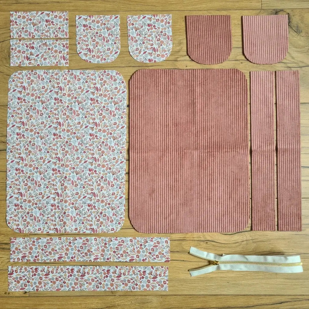 Kit "prêt à coudre" pour une trousse de toilette en velours côtelé au coloris rose avec une doublure en tissu à motif liberty. L'ensemble des pièces de tissu, déjà prédécoupées aux bonnes dimensions, sont disposées au sol. A gauche, sept pièces en tissu à motif liberty. A droite, cinq pièces en velours côtelé de couleur rose ainsi qu'une fermeture éclair blanche.