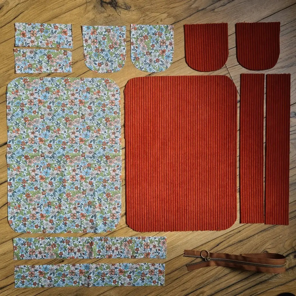 Kit "prêt à coudre" pour une trousse de toilette en velours côtelé au coloris terracotta avec une doublure en tissu à motif fleuri. L'ensemble des pièces de tissu, déjà prédécoupées aux bonnes dimensions, sont disposées au sol. A gauche, sept pièces en tissu à motif fleuri. A droite, cinq pièces en velours côtelé de couleur terracotta ainsi qu'une fermeture éclair marron.