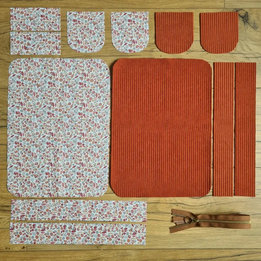 Kit "prêt à coudre" pour une trousse de toilette en velours côtelé au coloris terracotta avec une doublure en tissu à motif liberty. L'ensemble des pièces de tissu, déjà prédécoupées aux bonnes dimensions, sont disposées au sol. A gauche, sept pièces en tissu à motif liberty. A droite, cinq pièces en velours côtelé de couleur terracotta ainsi qu'une fermeture éclair marron.