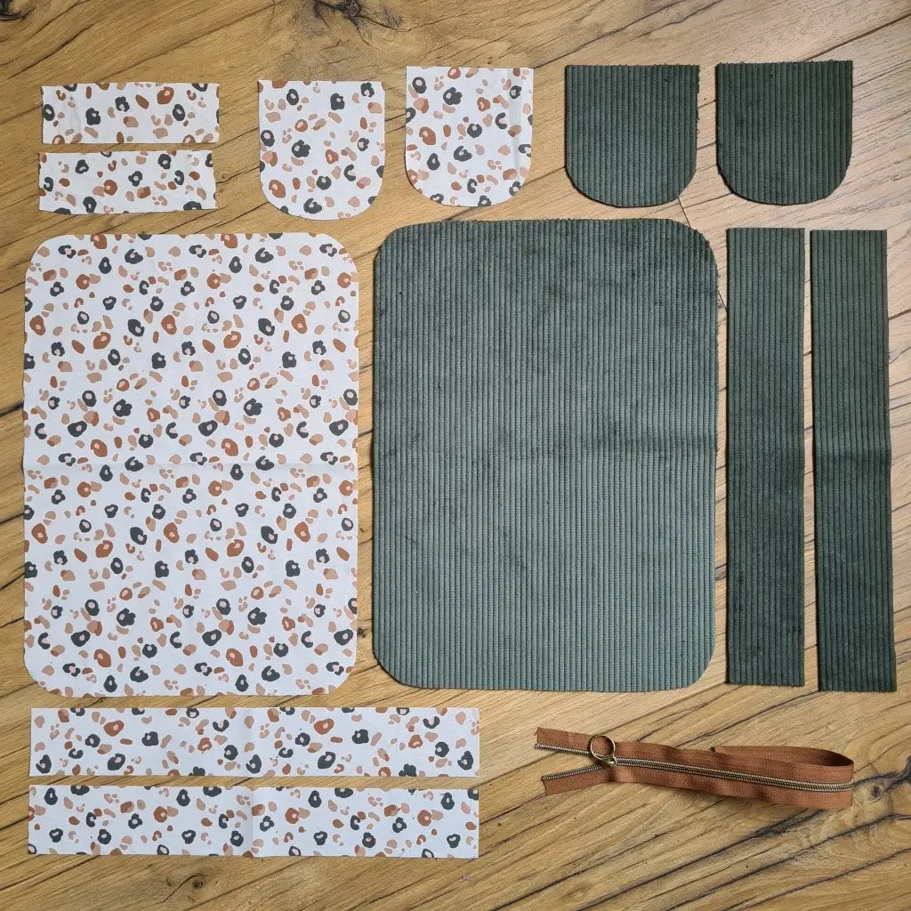 Kit "prêt à coudre" pour une trousse de toilette en velours côtelé au coloris vert avec une doublure en tissu à motif léopard. L'ensemble des pièces de tissu, déjà prédécoupées aux bonnes dimensions, sont disposées au sol. A gauche, sept pièces en tissu à motif léopard. A droite, cinq pièces en velours côtelé de couleur verte ainsi qu'une fermeture éclair marron.