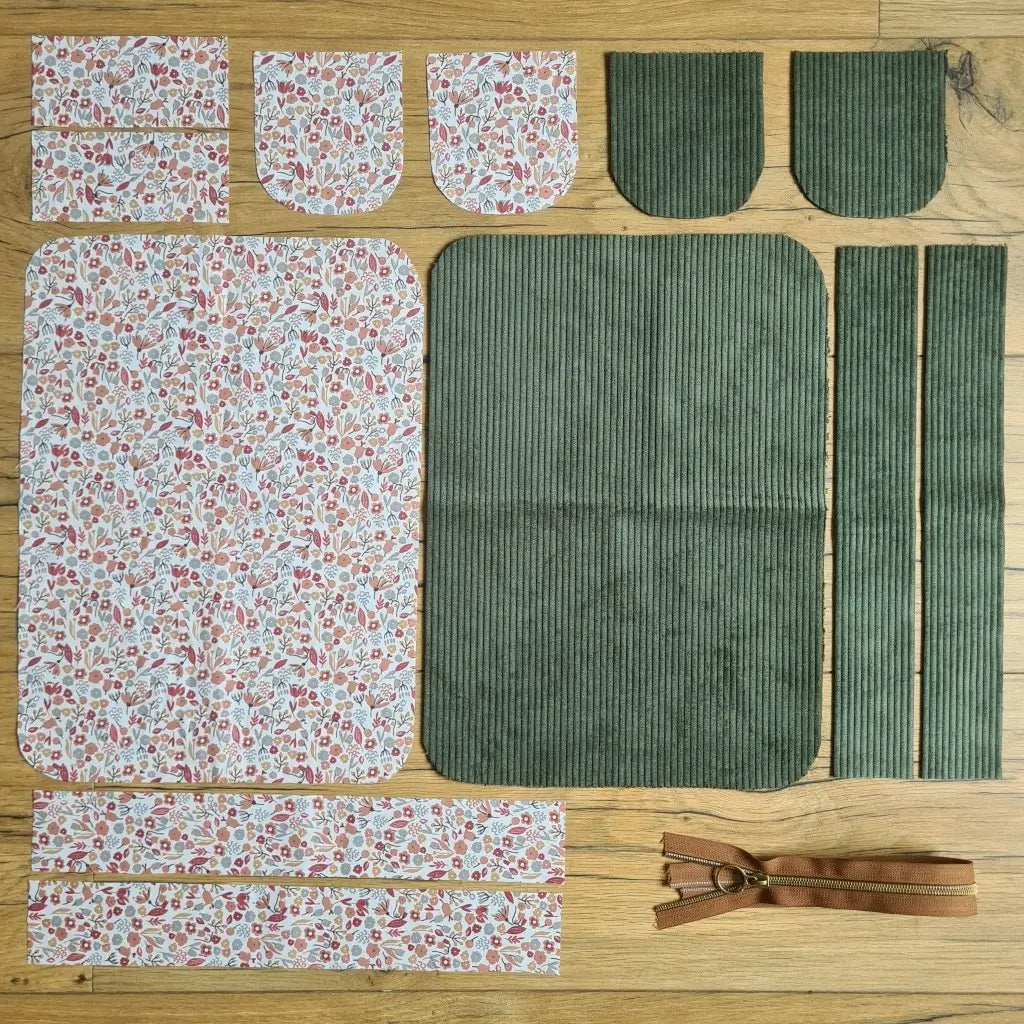 Kit "prêt à coudre" pour une trousse de toilette en velours côtelé au coloris vert avec une doublure en tissu à motif liberty. L'ensemble des pièces de tissu, déjà prédécoupées aux bonnes dimensions, sont disposées au sol. A gauche, sept pièces en tissu à motif liberty. A droite, cinq pièces en velours côtelé de couleur verte ainsi qu'une fermeture éclair marron.