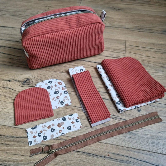 Kit "prêt à coudre" pour une trousse de toilette en velours côtelé de coloris terracotta, doublée avec un tissu intérieur de motif léopard. A l'arrière plan se trouve la trousse de toilette finie. Au premier plan sont disposées l'ensemble des pièces de tissu prédécoupées à la bonne dimension nécessaires pour la confection de cette trousse de toilette.