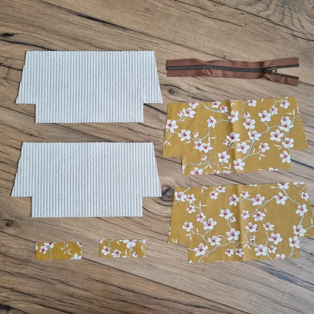 Kit prêt-à-coudre pour trousse d’école personnalisée comprenant deux pièces en velours côtelé beige, quatre coupons en coton enduit jaune à motif fleuri blanc et rose, une fermeture éclair marron et deux petites bandes assorties. Les éléments sont disposés à plat sur un sol en bois, prêts à être assemblés pour confectionner une trousse d'école zippée doublée, idéale pour la rentrée ou comme idée cadeau DIY.