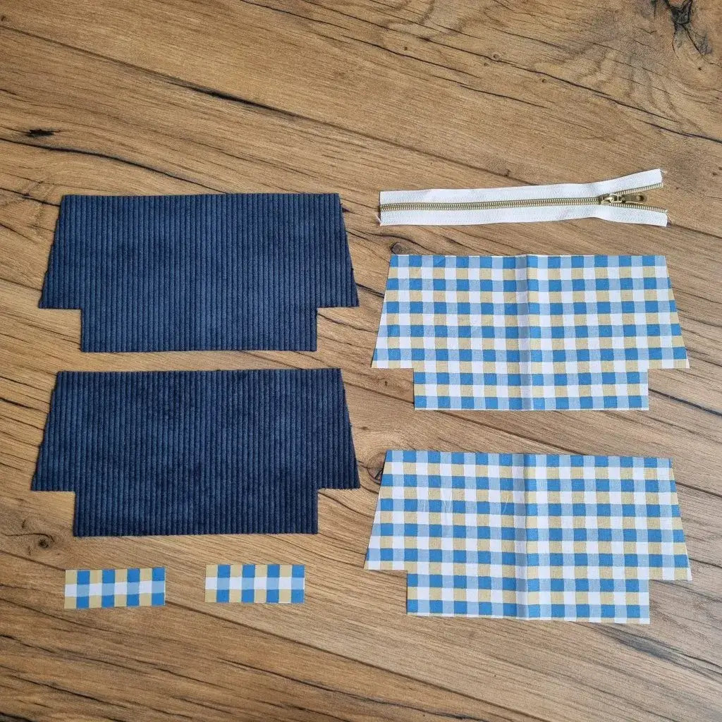 Kit prêt-à-coudre pour trousse d’école en velours côtelé bleu marine, composé de deux pièces extérieures, quatre coupons de doublure en coton enduit à carreaux bleu, blanc et ocre, deux petites bandes assorties et une fermeture éclair blanche avec zip doré. Tous les éléments sont présentés à plat sur un sol en bois, parfaits pour créer une trousse d'école personnalisée, robuste et tendance, idéale pour les fournitures scolaires.