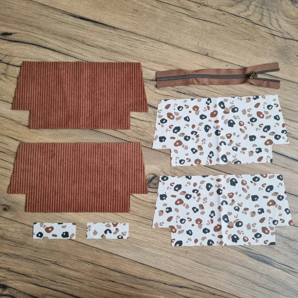 Kit prêt-à-coudre pour trousse d’école en velours côtelé marron, composé de deux pièces extérieures, quatre coupons en coton enduit imprimé léopard noir et brun sur fond blanc, deux petites bandes assorties et une fermeture éclair marron. Présentés à plat sur un sol en bois, ces éléments permettent de réaliser une trousse d'école zippée tendance et personnalisable, parfaite pour les amateurs de couture créative.