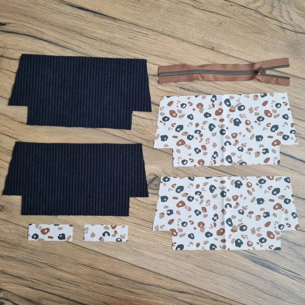 Kit de couture DIY pour trousse d’école en velours côtelé noir, comprenant deux pièces extérieures, quatre coupons en coton enduit à motif léopard noir et brun sur fond écru, deux petites bandes assorties et une fermeture éclair marron avec zip métallique. Les éléments sont disposés à plat sur un parquet en bois, prêts à être assemblés pour créer une trousse d'école personnalisée zippée, idéale pour une rentrée stylée ou un projet créatif fait main.