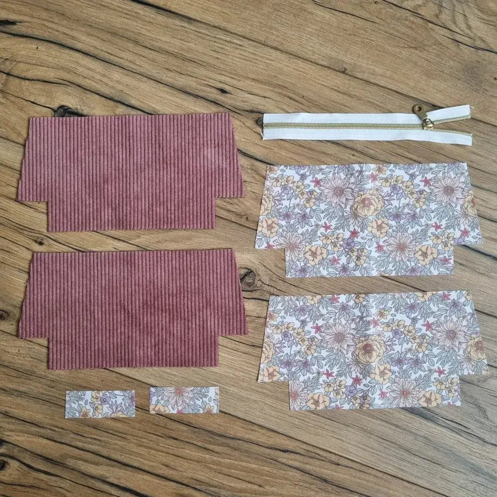 Kit prêt-à-coudre pour trousse d’école en velours côtelé rose, composé de deux pièces extérieures, quatre coupons en coton enduit imprimé floral aux tons beige, rouge et jaune, deux petites bandes assorties et une fermeture éclair blanche à zip doré. Les éléments sont présentés à plat sur un sol en bois, parfaits pour réaliser une trousse d'école personnalisée zippée avec doublure, idéale pour un projet couture créatif et pratique.