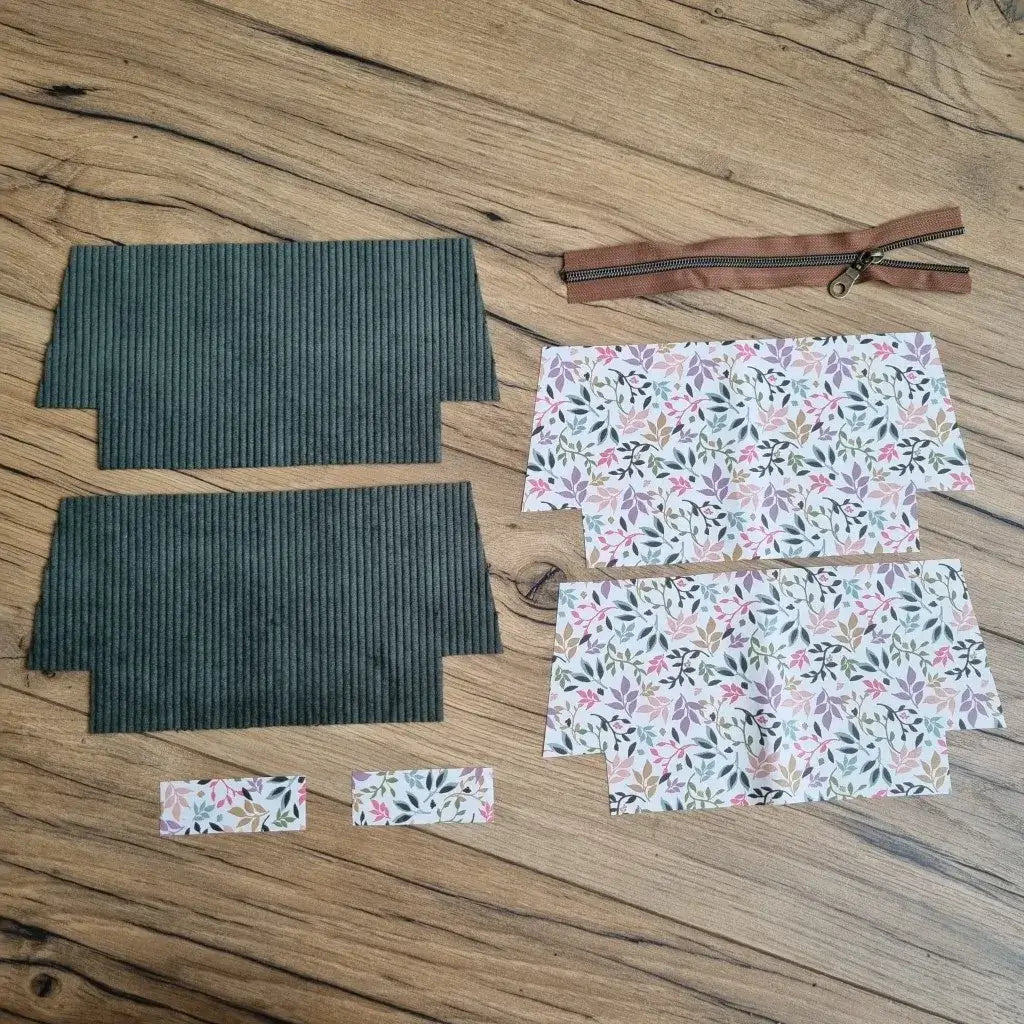 Kit prêt-à-coudre pour trousse d’école en velours côtelé vert, comprenant deux pièces extérieures, quatre coupons de doublure en coton enduit à motif fleuri pastel, deux petites bandes assorties et une fermeture éclair marron avec zip métallique. Tous les éléments sont étalés sur un sol en bois, prêts à être assemblés pour créer une trousse d'école zippée personnalisée, parfaite pour un projet couture simple et utile.