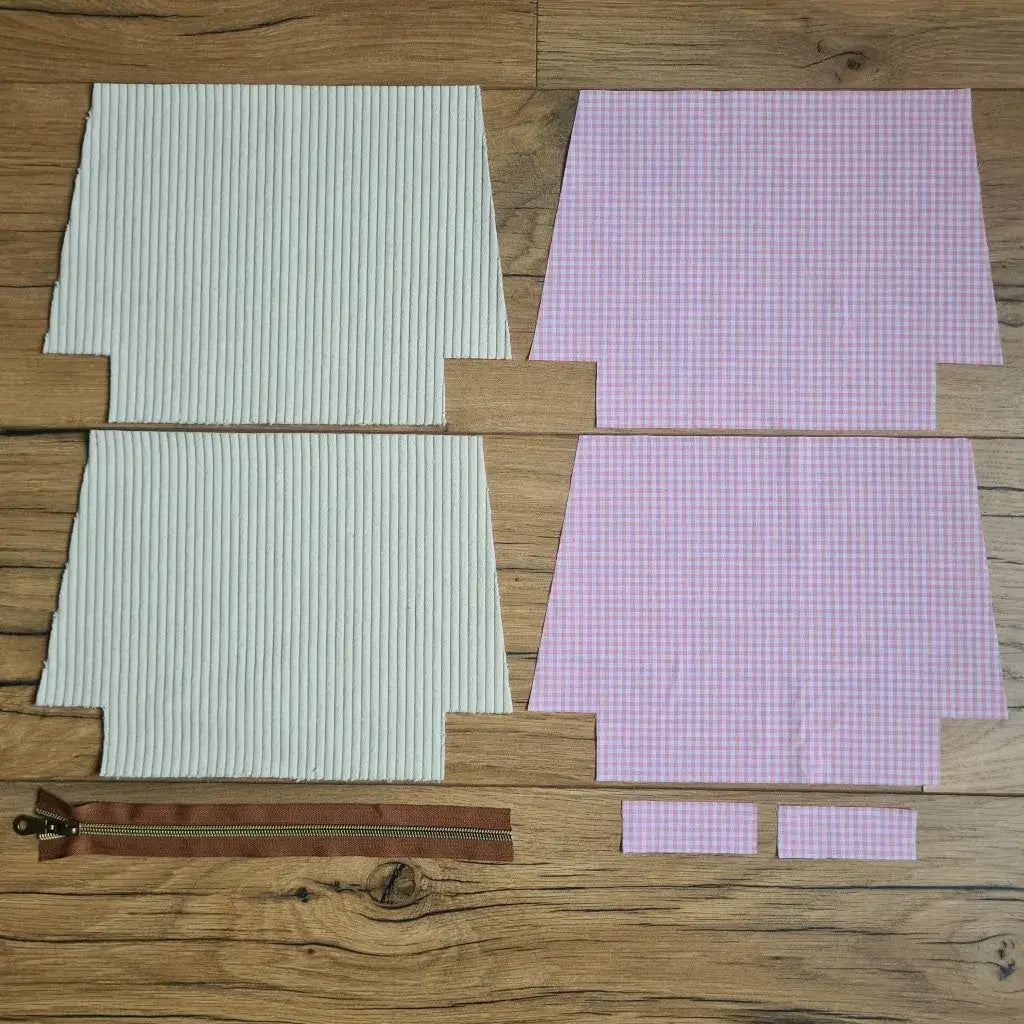 Kit "prêt à coudre" pour une trousse zippée en velours côtelé au coloris beige avec une doublure en tissu à motif carreaux roses. L'ensemble des pièces de tissu, déjà prédécoupées aux bonnes dimensions, sont disposées au sol. A gauche, deux pièces identiques en velours côtelé beige ainsi qu'une fermeture éclair marron. A droite, quatre pièces en tissu imprimé à motif carreaux roses.