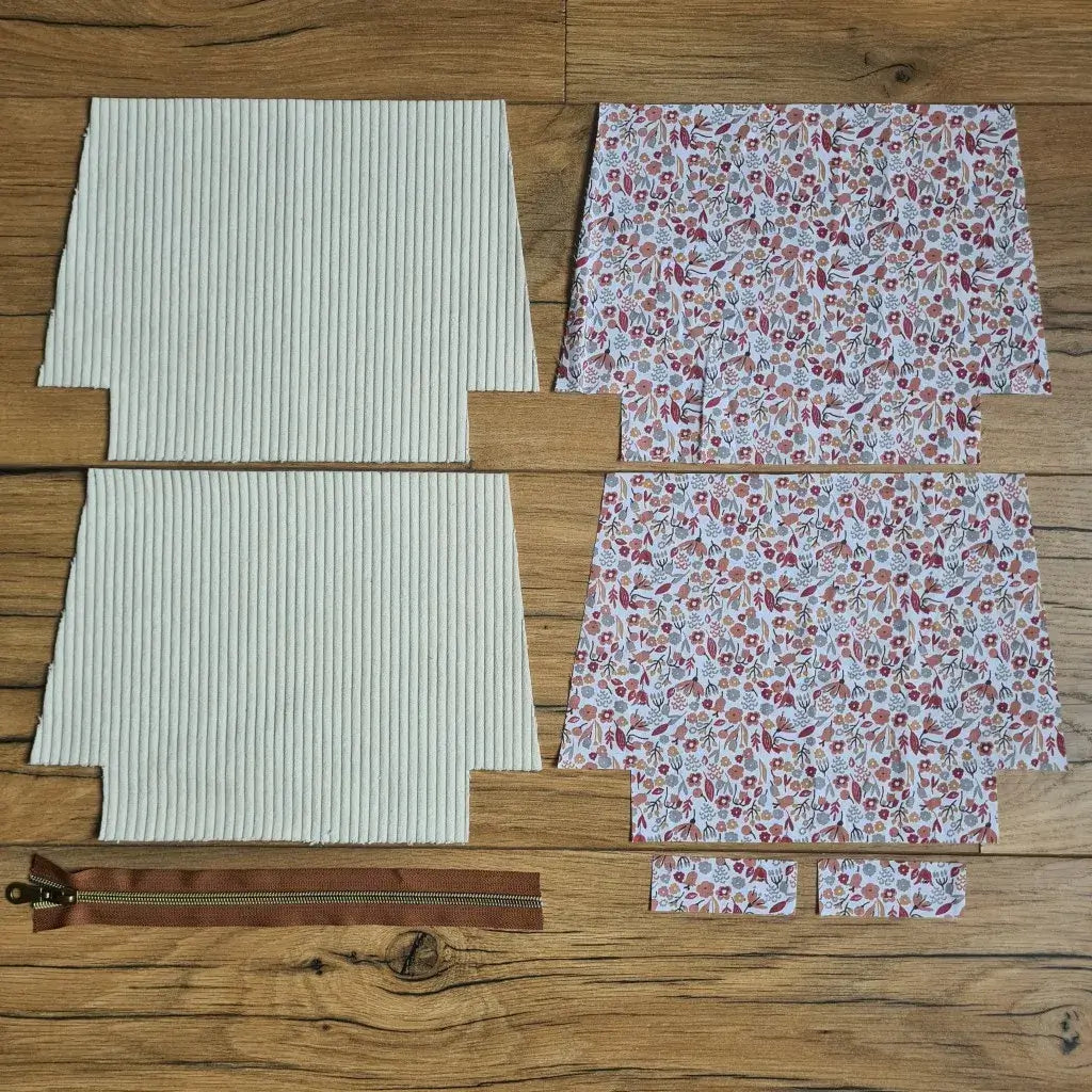 Kit "prêt à coudre" pour une trousse zippée en velours côtelé au coloris beige avec une doublure en tissu à motif liberty. L'ensemble des pièces de tissu, déjà prédécoupées aux bonnes dimensions, sont disposées au sol. A gauche, deux pièces identiques en velours côtelé beige ainsi qu'une fermeture éclair marron. A droite, quatre pièces en tissu imprimé à motif liberty.