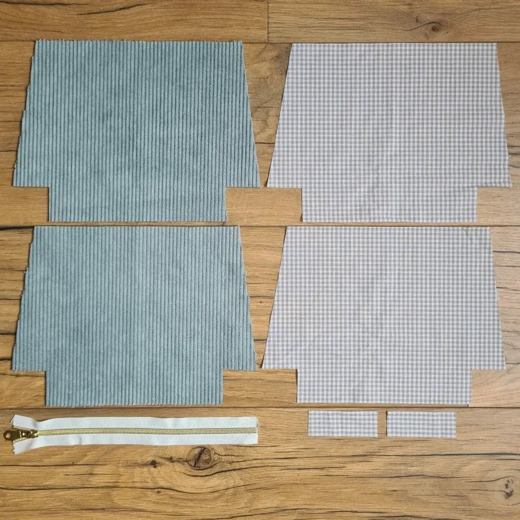 Kit "prêt à coudre" pour une trousse zippée en velours côtelé au coloris bleu clair avec une doublure en tissu à motif carreaux beiges. L'ensemble des pièces de tissu, déjà prédécoupées aux bonnes dimensions, sont disposées au sol. A gauche, deux pièces identiques en velours côtelé bleu clair ainsi qu'une fermeture éclair blanche. A droite, quatre pièces en tissu imprimé à motif carreaux beiges.