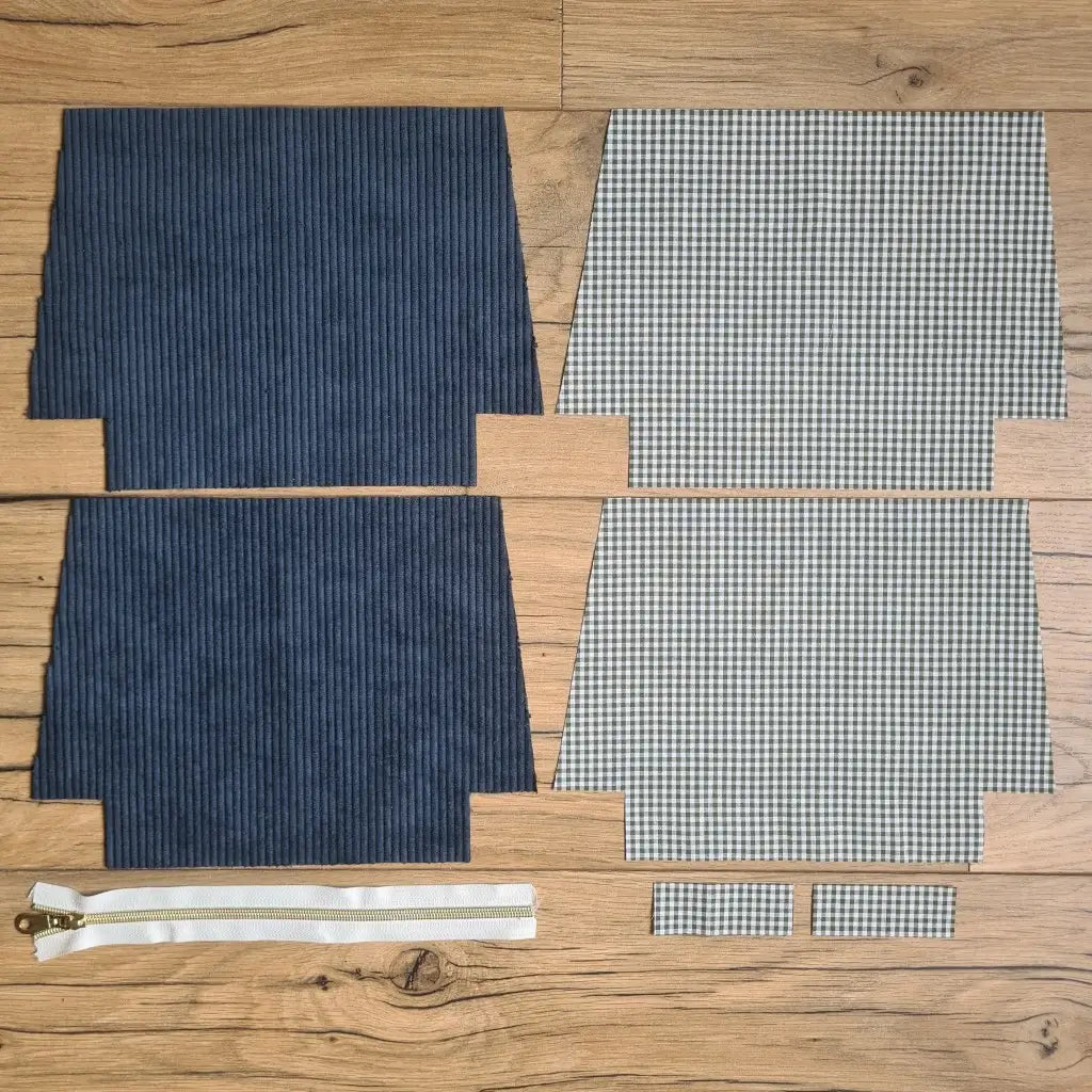 Kit "prêt à coudre" pour une trousse zippée en velours côtelé au coloris bleu foncé avec une doublure en tissu à motif carreaux verts. L'ensemble des pièces de tissu, déjà prédécoupées aux bonnes dimensions, sont disposées au sol. A gauche, deux pièces identiques en velours côtelé bleu foncé ainsi qu'une fermeture éclair blanche. A droite, quatre pièces en tissu imprimé à motif carreaux verts.