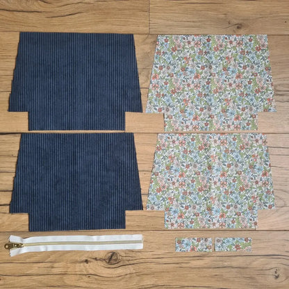 Kit "prêt à coudre" pour une trousse zippée en velours côtelé au coloris bleu foncé avec une doublure en tissu à motif fleuri. L'ensemble des pièces de tissu, déjà prédécoupées aux bonnes dimensions, sont disposées au sol. A gauche, deux pièces identiques en velours côtelé bleu foncé ainsi qu'une fermeture éclair blanche. A droite, quatre pièces en tissu imprimé à motif fleuri.
