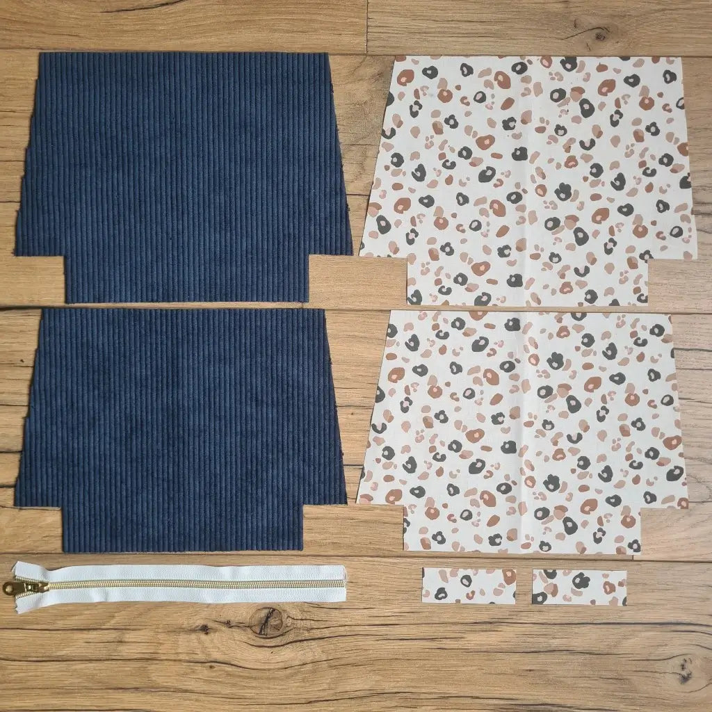 Kit "prêt à coudre" pour une trousse zippée en velours côtelé au coloris bleu foncé avec une doublure en tissu à motif léopard. L'ensemble des pièces de tissu, déjà prédécoupées aux bonnes dimensions, sont disposées au sol. A gauche, deux pièces identiques en velours côtelé bleu foncé ainsi qu'une fermeture éclair blanche. A droite, quatre pièces en tissu imprimé à motif léopard.