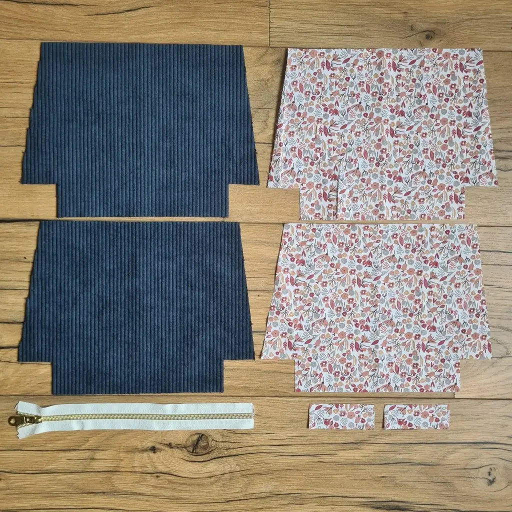 Kit "prêt à coudre" pour une trousse zippée en velours côtelé au coloris bleu foncé avec une doublure en tissu à motif liberty. L'ensemble des pièces de tissu, déjà prédécoupées aux bonnes dimensions, sont disposées au sol. A gauche, deux pièces identiques en velours côtelé bleu foncé ainsi qu'une fermeture éclair blanche. A droite, quatre pièces en tissu imprimé à motif liberty.