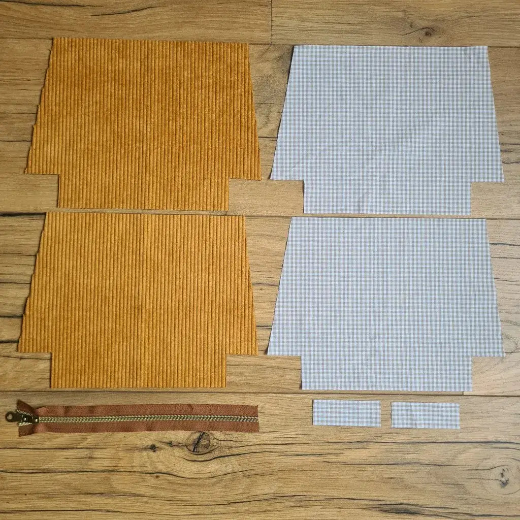 Kit "prêt à coudre" pour une trousse zippée en velours côtelé au coloris jaune avec une doublure en tissu à motif carreaux beiges. L'ensemble des pièces de tissu, déjà prédécoupées aux bonnes dimensions, sont disposées au sol. A gauche, deux pièces identiques en velours côtelé jaune ainsi qu'une fermeture éclair marron. A droite, quatre pièces en tissu imprimé à motif carreaux beiges.