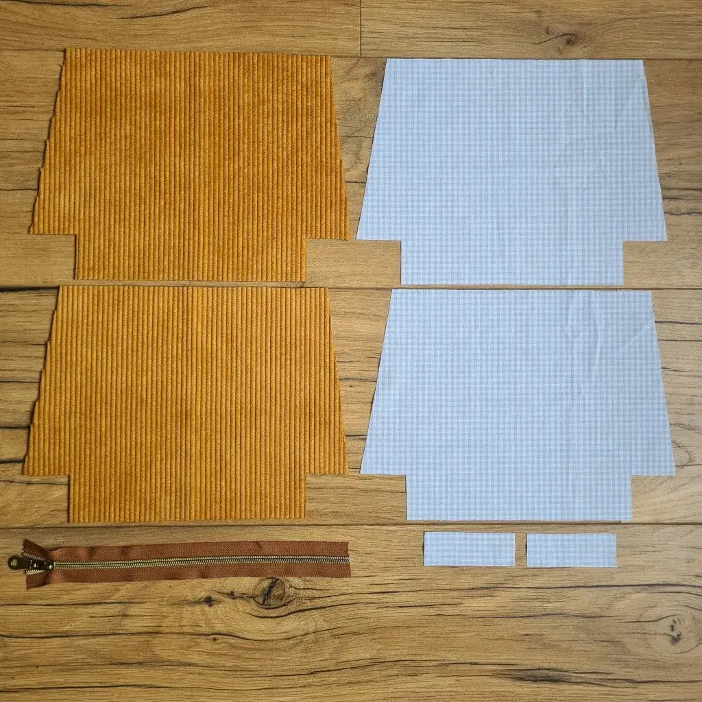 Kit "prêt à coudre" pour une trousse zippée en velours côtelé au coloris jaune avec une doublure en tissu à motif carreaux bleus. L'ensemble des pièces de tissu, déjà prédécoupées aux bonnes dimensions, sont disposées au sol. A gauche, deux pièces identiques en velours côtelé jaune ainsi qu'une fermeture éclair marron. A droite, quatre pièces en tissu imprimé à motif carreaux bleus.