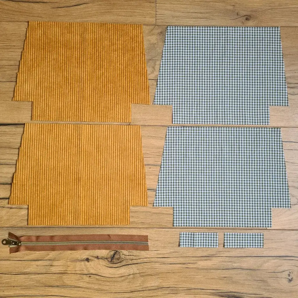 Kit "prêt à coudre" pour une trousse zippée en velours côtelé au coloris jaune avec une doublure en tissu à motif carreaux verts. L'ensemble des pièces de tissu, déjà prédécoupées aux bonnes dimensions, sont disposées au sol. A gauche, deux pièces identiques en velours côtelé jaune ainsi qu'une fermeture éclair marron. A droite, quatre pièces en tissu imprimé à motif carreaux verts.