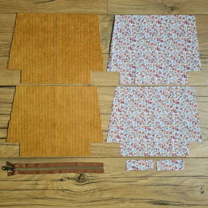Kit "prêt à coudre" pour une trousse zippée en velours côtelé au coloris jaune avec une doublure en tissu à motif liberty. L'ensemble des pièces de tissu, déjà prédécoupées aux bonnes dimensions, sont disposées au sol. A gauche, deux pièces identiques en velours côtelé jaune ainsi qu'une fermeture éclair marron. A droite, quatre pièces en tissu imprimé à motif liberty.