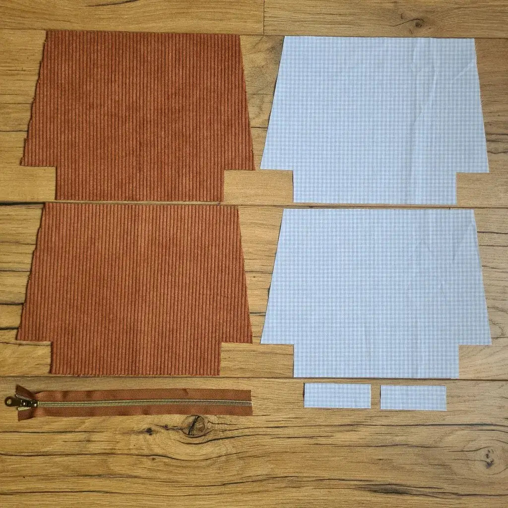 Kit "prêt à coudre" pour une trousse zippée en velours côtelé au coloris marron avec une doublure en tissu à motif carreaux bleus. L'ensemble des pièces de tissu, déjà prédécoupées aux bonnes dimensions, sont disposées au sol. A gauche, deux pièces identiques en velours côtelé marron ainsi qu'une fermeture éclair marron. A droite, quatre pièces en tissu imprimé à motif carreaux bleus.