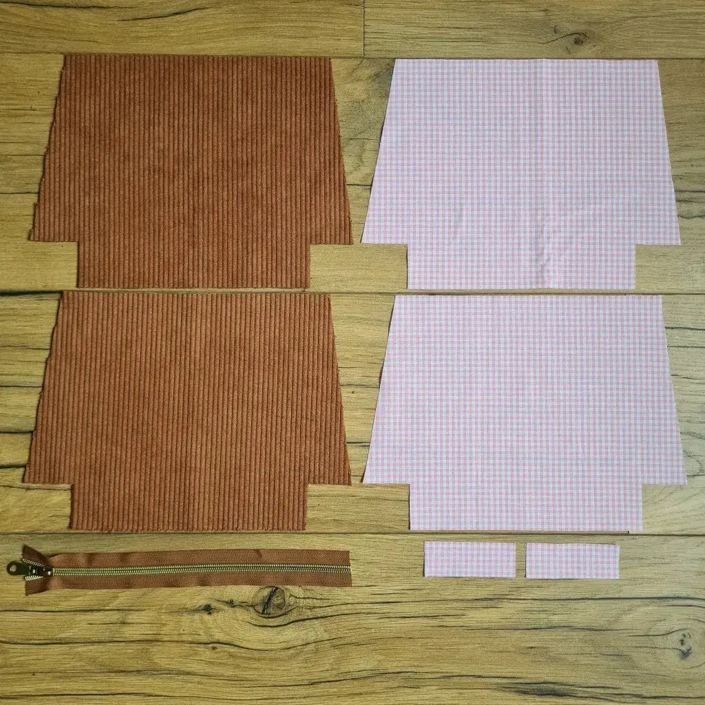 Kit "prêt à coudre" pour une trousse zippée en velours côtelé au coloris marron avec une doublure en tissu à motif carreaux roses. L'ensemble des pièces de tissu, déjà prédécoupées aux bonnes dimensions, sont disposées au sol. A gauche, deux pièces identiques en velours côtelé marron ainsi qu'une fermeture éclair marron. A droite, quatre pièces en tissu imprimé à motif carreaux roses.