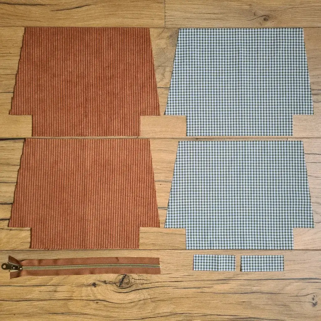 Kit "prêt à coudre" pour une trousse zippée en velours côtelé au coloris marron avec une doublure en tissu à motif carreaux verts. L'ensemble des pièces de tissu, déjà prédécoupées aux bonnes dimensions, sont disposées au sol. A gauche, deux pièces identiques en velours côtelé marron ainsi qu'une fermeture éclair marron. A droite, quatre pièces en tissu imprimé à motif carreaux verts.