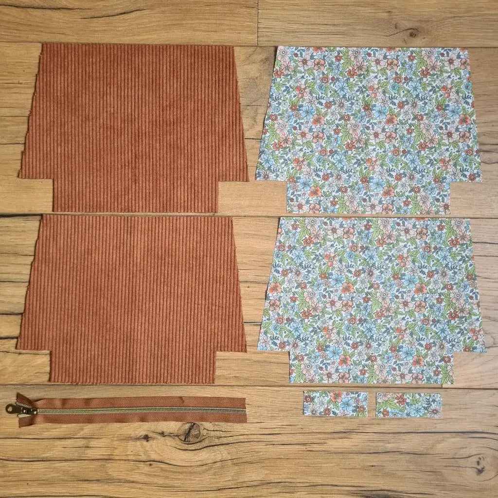 Kit "prêt à coudre" pour une trousse zippée en velours côtelé au coloris marron avec une doublure en tissu à motif fleuri. L'ensemble des pièces de tissu, déjà prédécoupées aux bonnes dimensions, sont disposées au sol. A gauche, deux pièces identiques en velours côtelé marron ainsi qu'une fermeture éclair marron. A droite, quatre pièces en tissu imprimé à motif fleuri.