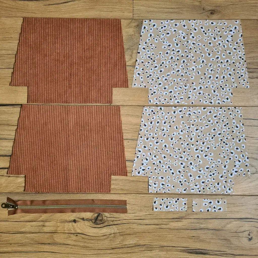 Kit "prêt à coudre" pour une trousse zippée en velours côtelé au coloris marron avec une doublure en tissu à motif léopard. L'ensemble des pièces de tissu, déjà prédécoupées aux bonnes dimensions, sont disposées au sol. A gauche, deux pièces identiques en velours côtelé marron ainsi qu'une fermeture éclair marron. A droite, quatre pièces en tissu imprimé à motif léopard.