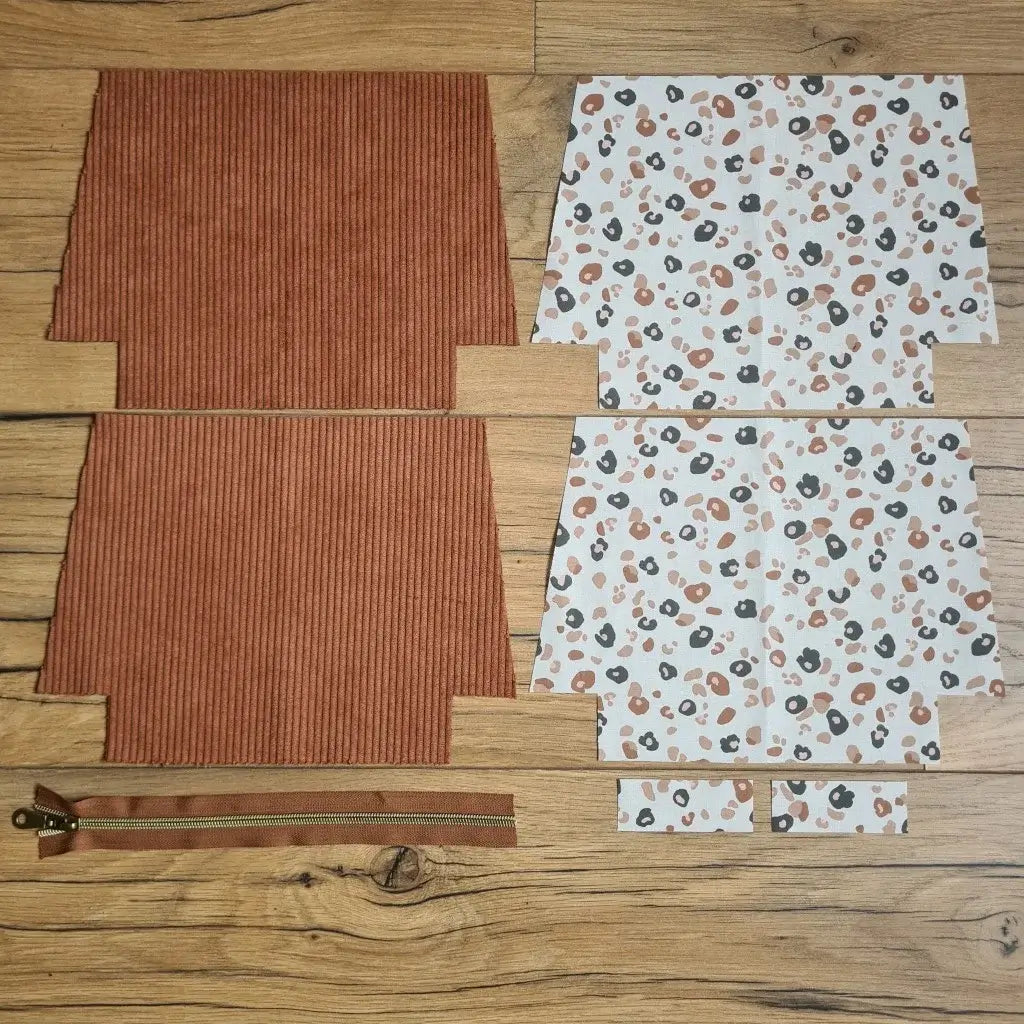 Kit "prêt à coudre" pour une trousse zippée en velours côtelé au coloris marron avec une doublure en tissu à motif léopard. L'ensemble des pièces de tissu, déjà prédécoupées aux bonnes dimensions, sont disposées au sol. A gauche, deux pièces identiques en velours côtelé marron ainsi qu'une fermeture éclair marron. A droite, quatre pièces en tissu imprimé à motif léopard.