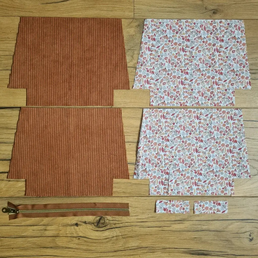 Kit "prêt à coudre" pour une trousse zippée en velours côtelé au coloris marron avec une doublure en tissu à motif liberty. L'ensemble des pièces de tissu, déjà prédécoupées aux bonnes dimensions, sont disposées au sol. A gauche, deux pièces identiques en velours côtelé marron ainsi qu'une fermeture éclair marron. A droite, quatre pièces en tissu imprimé à motif liberty.